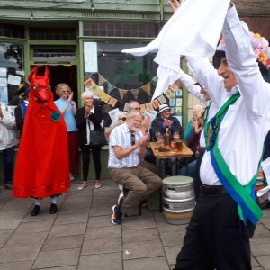 Morris Dancing outside a small pub in a converted high street shop - a Morris Dancer holds handkerchiefs aloft, and by the pub doorway a Morris animal with a red fox's head and a long red cape - called the Beast - looks on.